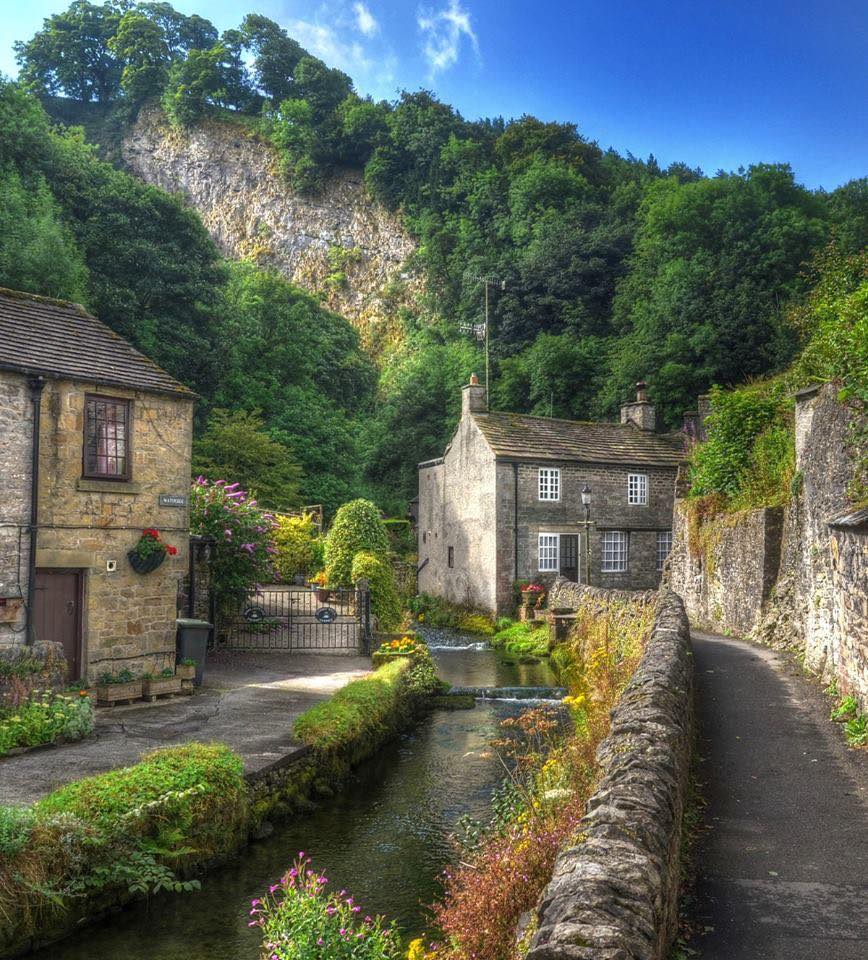 Stone cottages beside a stream and lane beneath a wooded cliff in Castleton, Peak District