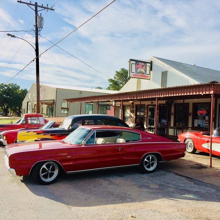 Red vintage muscle car parked at The Hill restaurant storefront with other classic cars visible