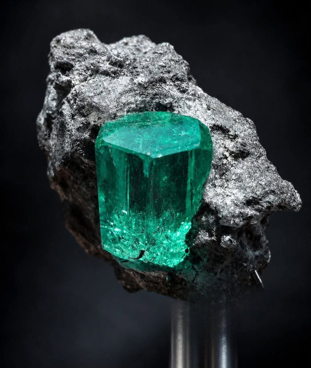 Bright green emerald crystal growing from a dark rock matrix on a display stand