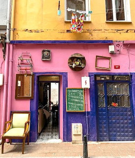 Colorful building exterior with yellow, pink, purple, and blue painted walls, door, chair, and sidewalk chalkboard