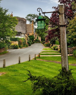 Quiet village street with thatched cottages, green lawns, and a vintage lamp post in the English Cotswolds
