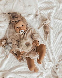 Baby with pacifier in beige outfit lying on white bedding surrounded by toys