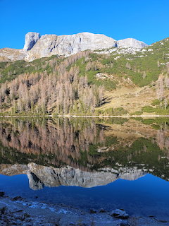 Rocky mountain and sparse trees reflected in a calm lake beneath a clear blue sky