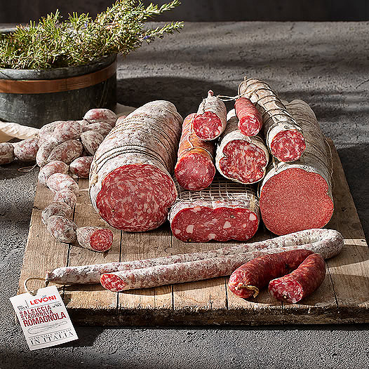 Assorted Italian salami and cured meats displayed on burlap with herbs and a wooden board