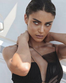 Dark-haired woman with green eyes in black sheer top posing with hands behind neck
