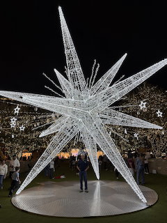 Large white wire-frame star sculpture lit up at night with people standing nearby under string lights