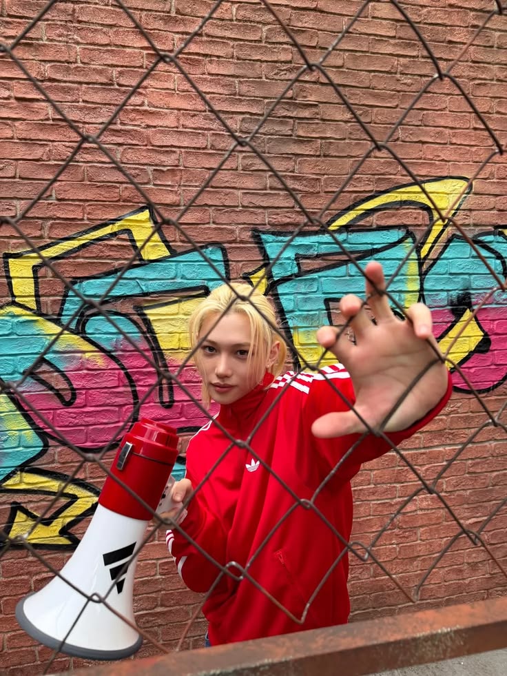 Young man in red Adidas jacket holding a megaphone, posing by a graffiti wall behind a chain-link fence.
