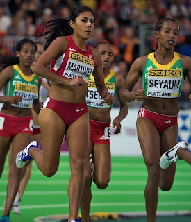 Female athletes sprinting in an indoor track competition, wearing red and green uniforms