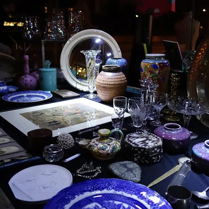 Antique table display with pottery, glassware, plates, and framed artwork in a dim interior
