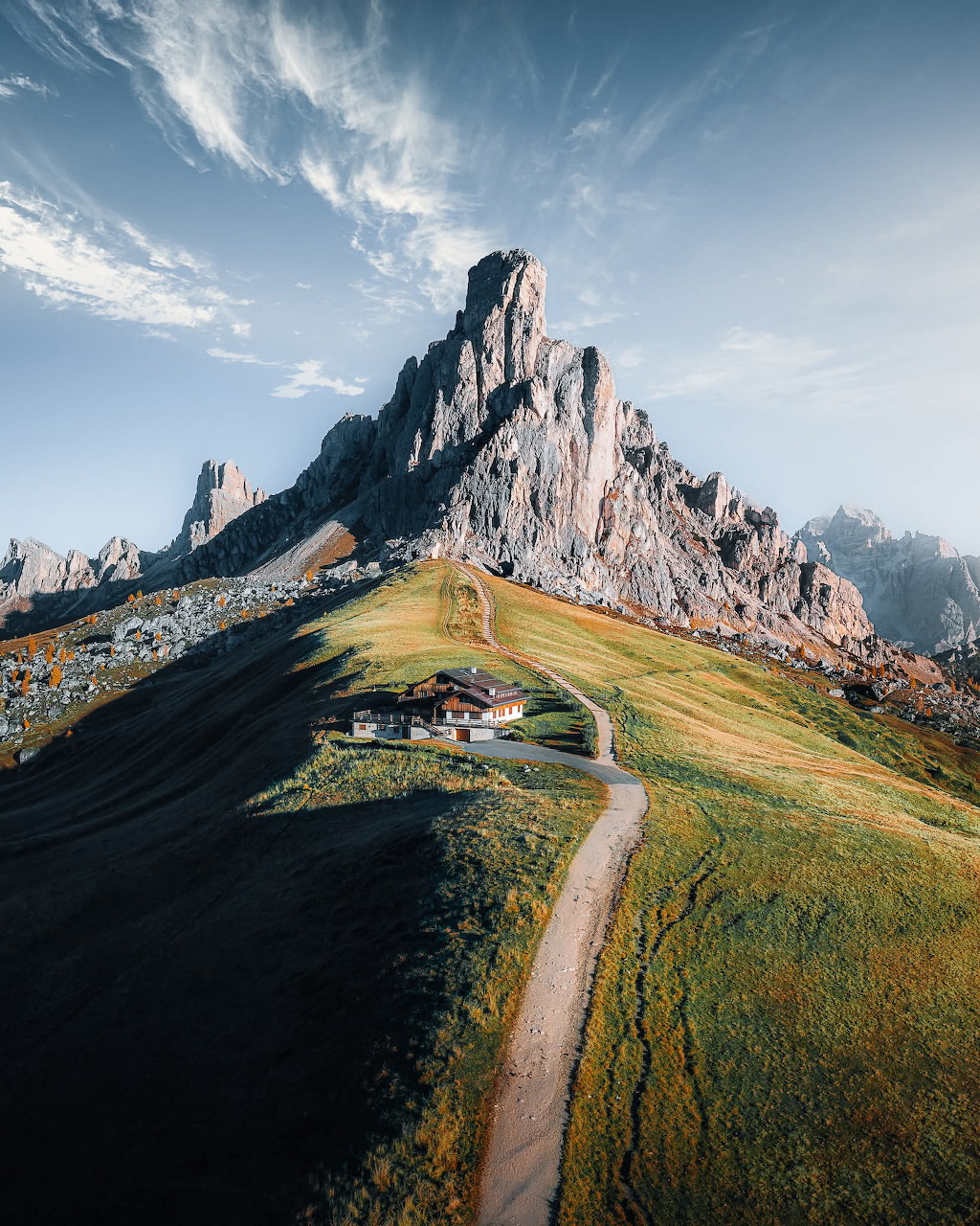 Lone alpine chalet on green hillside below jagged Dolomite rock peaks with winding path