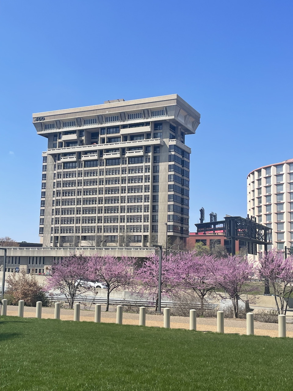 Brutalist office tower with protruding top floor, cherry trees in bloom, and grass lawn in foreground