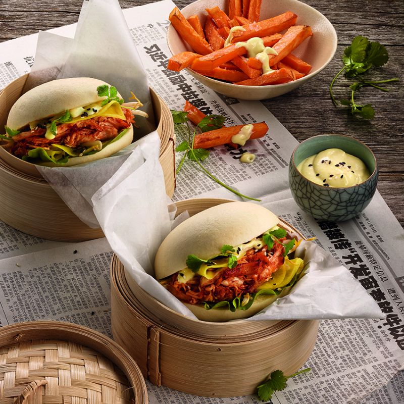 Two bao buns with pulled chicken, lettuce, and vegetables served in bamboo steamers with carrot sticks and corn