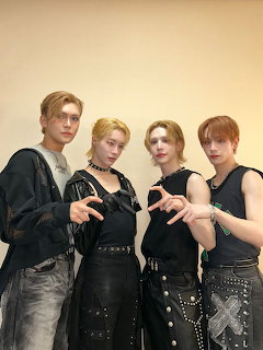 Four young men in black clothing posing together with hand gestures against a beige wall