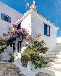 White Greek house with blue shutters and pink bougainvillea climbing the walls under a blue sky