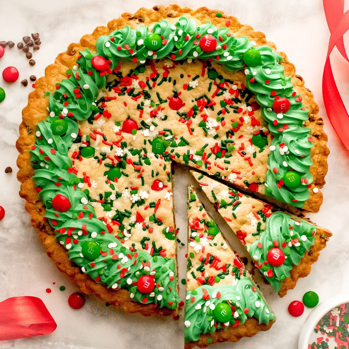 Round Christmas cookie cake with green icing wreath border, red candies, and sprinkles, with one slice removed
