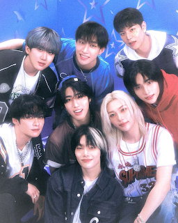 Eight K-pop group members posing closely together with colorful hair against a blue backdrop with stars