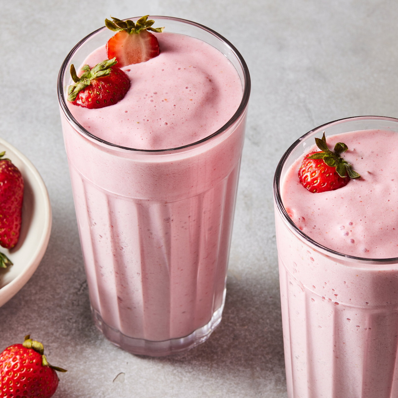 Two glasses of pink strawberry smoothie topped with fresh strawberries on gray counter