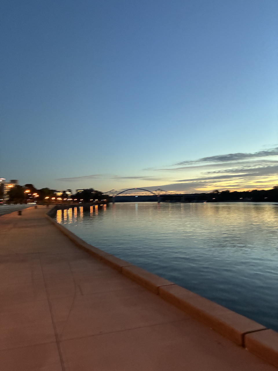 Riverside walkway at sunset with an arch bridge and city lights reflecting on calm water
