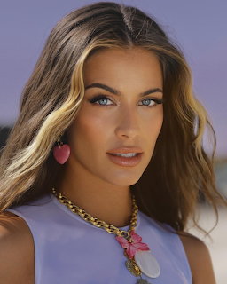 Portrait of woman with blonde hair and blue eyes wearing purple top and pink jewelry