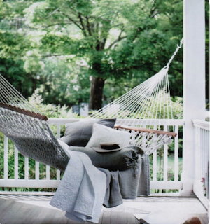 Woven hammock on a porch beside a cushioned chair with a throw blanket and leafy trees beyond