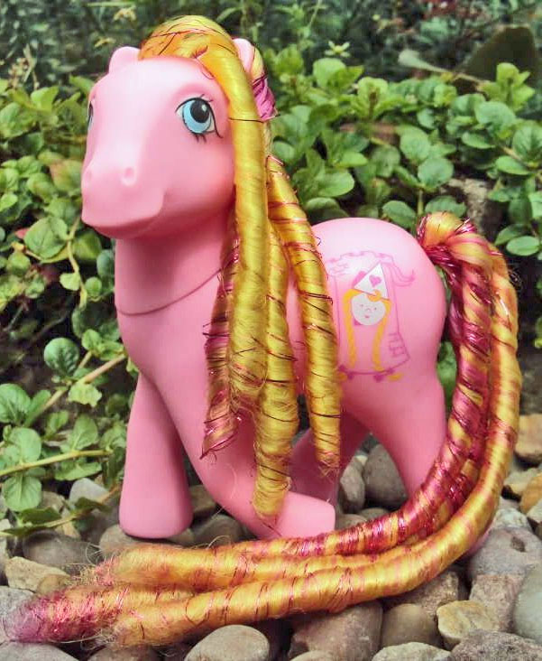 Pink toy pony with long golden-orange hair standing on pebbles among green leaves