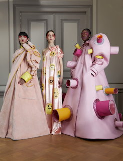 Three models in voluminous designer coats with colorful sphere decorations standing in elegant room