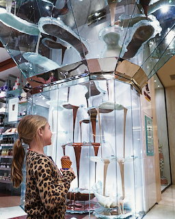 Young girl in leopard print top watching a tall chocolate fountain display behind glass