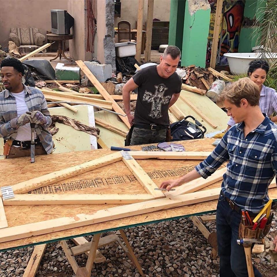 Three people assembling a large wooden frame outdoors with tools and lumber around them