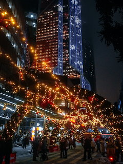 City street at night lined with glowing Christmas lights and market stalls beneath tall skyscrapers