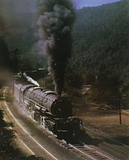 Steam locomotive emitting black smoke on curved tracks through a woded mountain valley