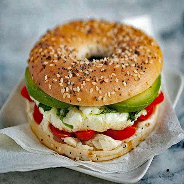 Seeded bagel sandwich with avocado, tomato, greens, and creamy cheese on a plate.