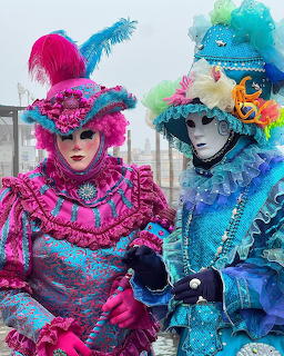Two people in elaborate pink and blue Venetian carnival costumes with ornate masks and feathered headdresses
