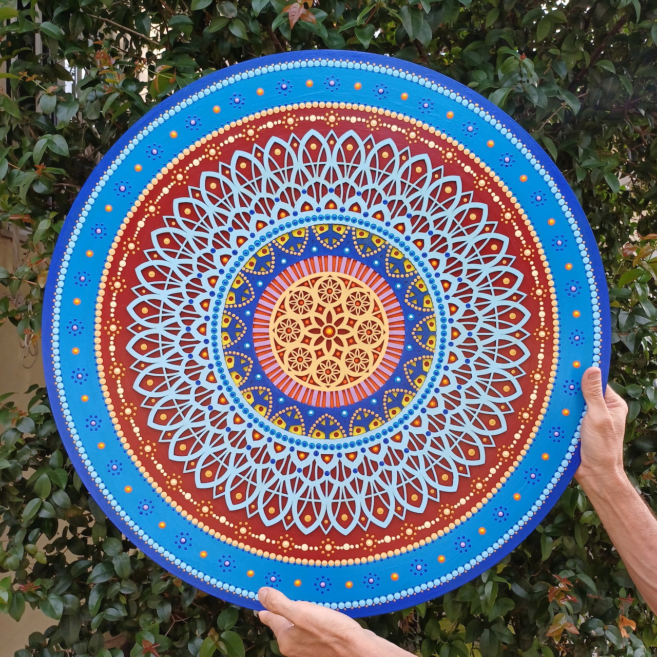 Hands holding circular mandala art with intricate geometric patterns in gold, red, and blue against green foliage