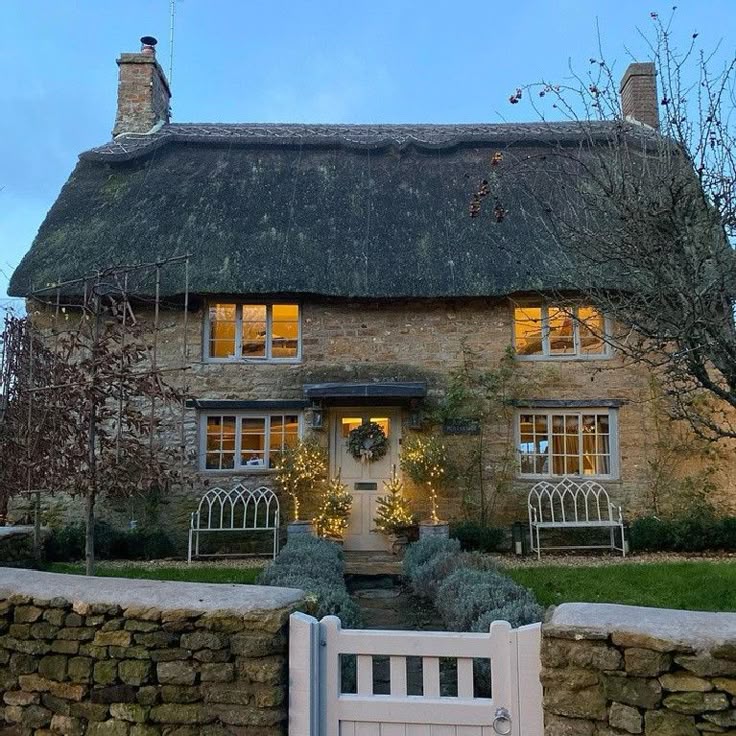 Stone cottage with a thatched roof, lit windows, white gate, and stone wall at dusk