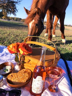 Brown horse sniffing picnic basket with wine, roses, and food on checkered blanket outdoors
