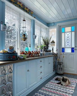 Blue country kitchen interior with lace curtains, decorative items on windowsill, and a Siamese cat on a rug