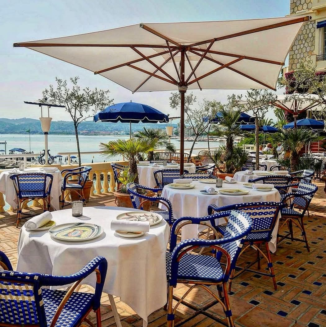 Outdoor restaurant terrace with white-clothed tables, blue chairs, and umbrellas overlooking the sea
