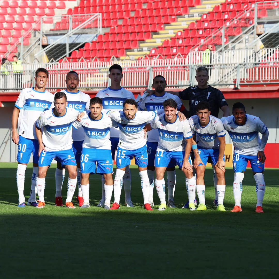 Soccer team in white and blue uniforms posing on a stadium field in front of red seats