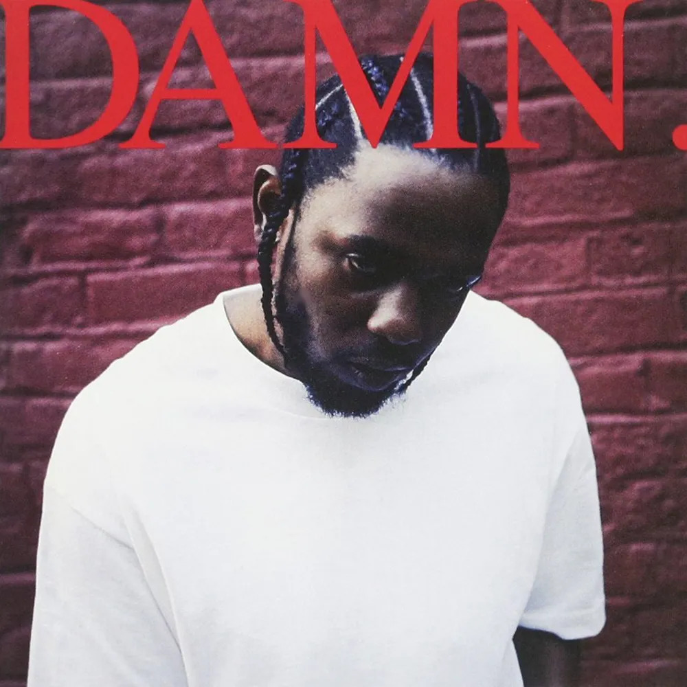 Kendrick Lamar in white t-shirt looking down against red brick wall with DAMN text above