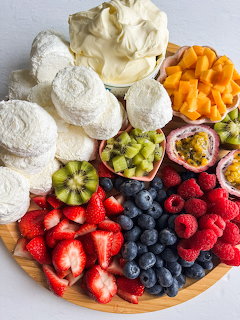 Wooden serving board with assorted fresh fruits, white meringue cookies, and whipped cream on light background