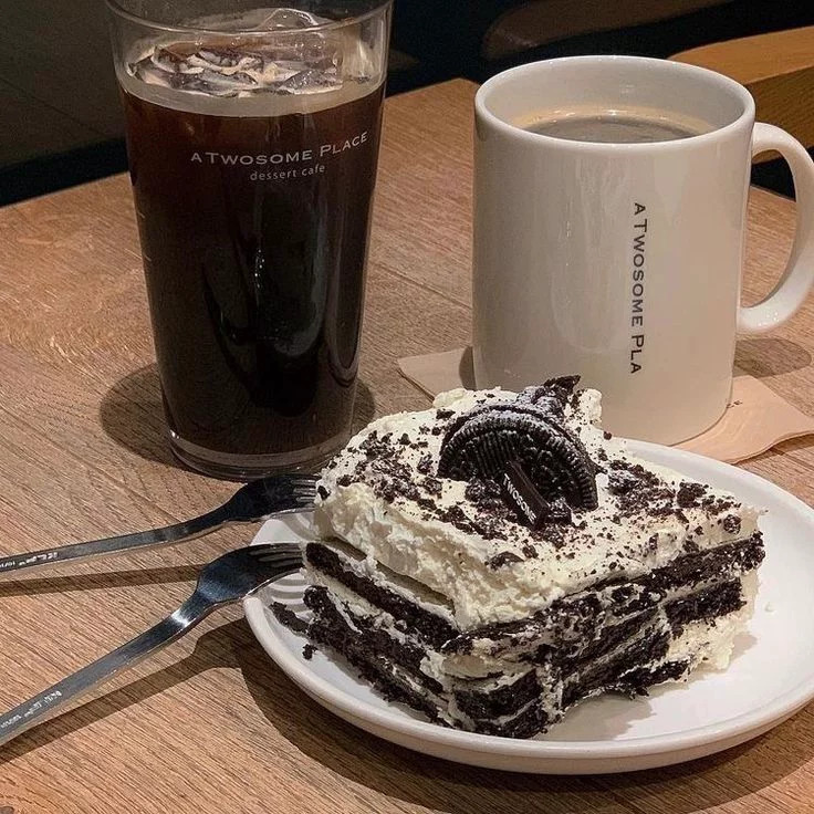 Slice of Oreo cream cake with iced coffee and a hot coffee mug on a wooden cafe table