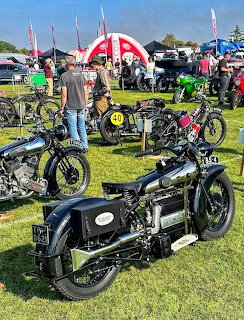 Several vintage black motorcycles displayed on grass at an outdoor motorcycle show with people and tents