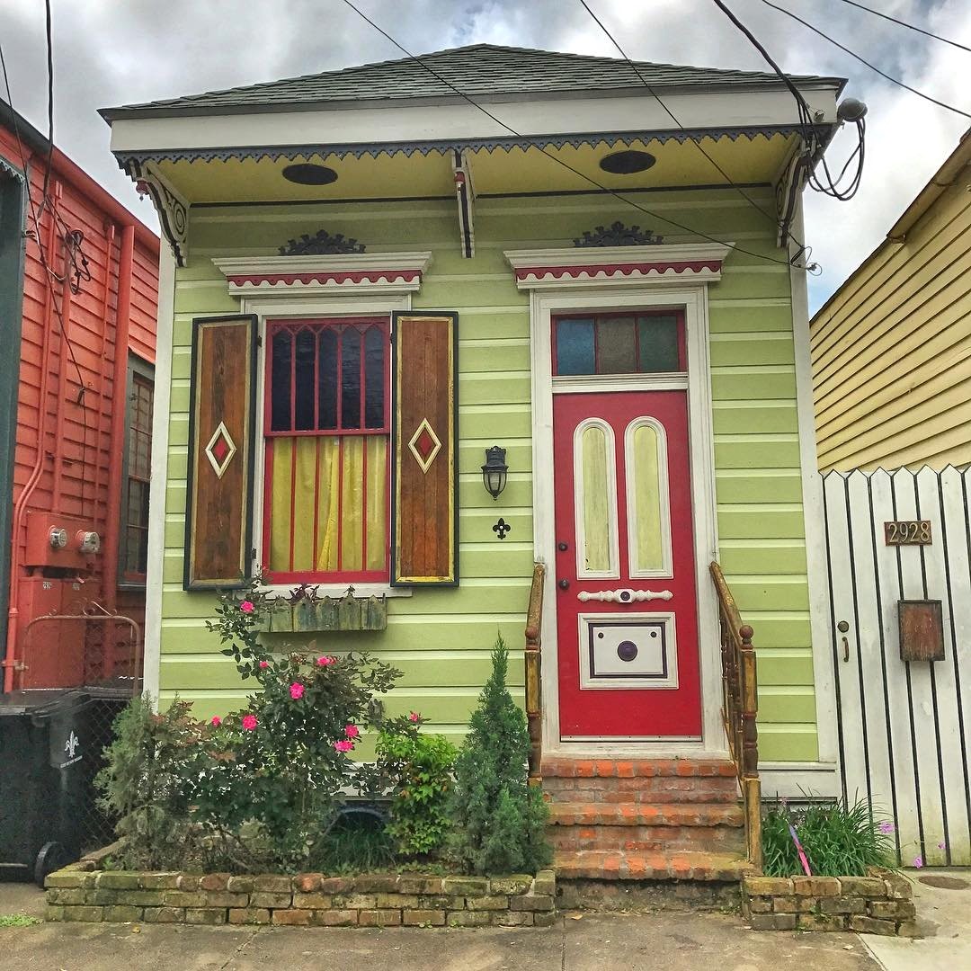 Olive green shotgun house with red door between colorful neighboring homes on urban street