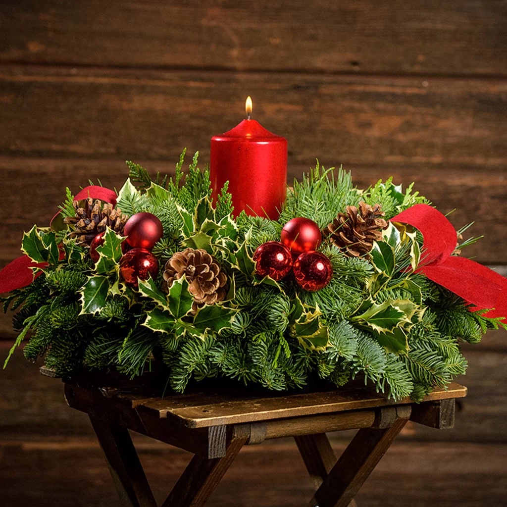 Christmas centerpiece with lit red candle surrounded by evergreen branches, pine cones, and red ornaments on wooden table