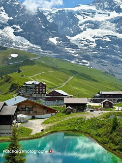 Swiss alpine village with a turquoise pond, green hills, and snow-covered mountains