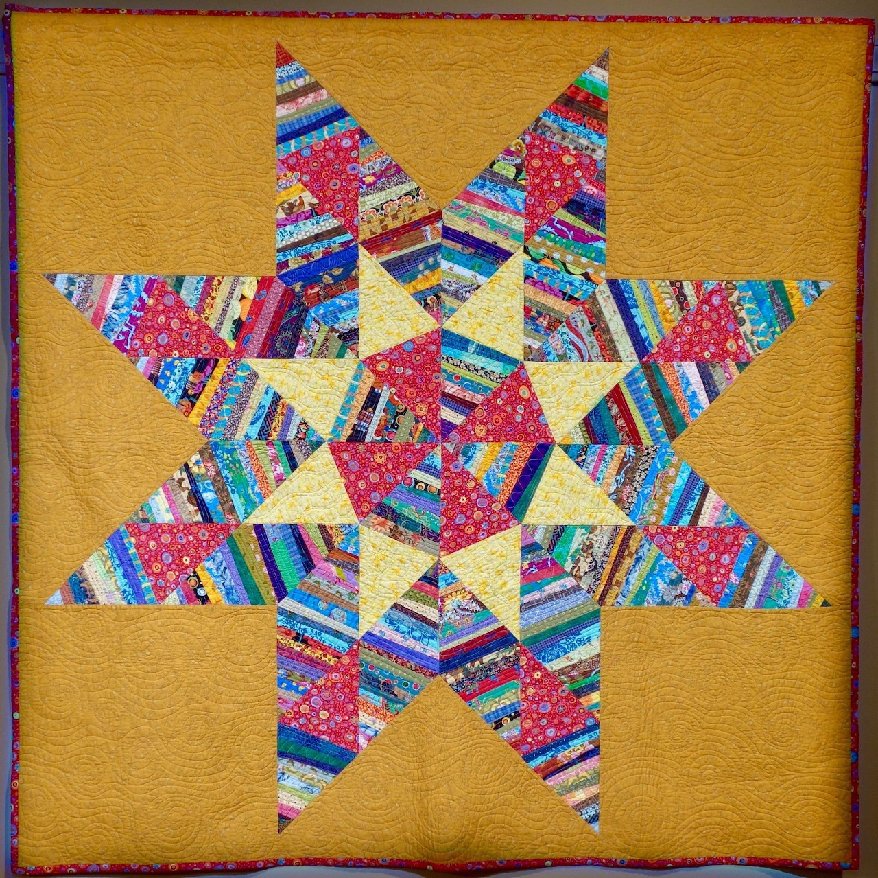 Geometric star quilt with multicolored patchwork strips radiating from center on gold fabric