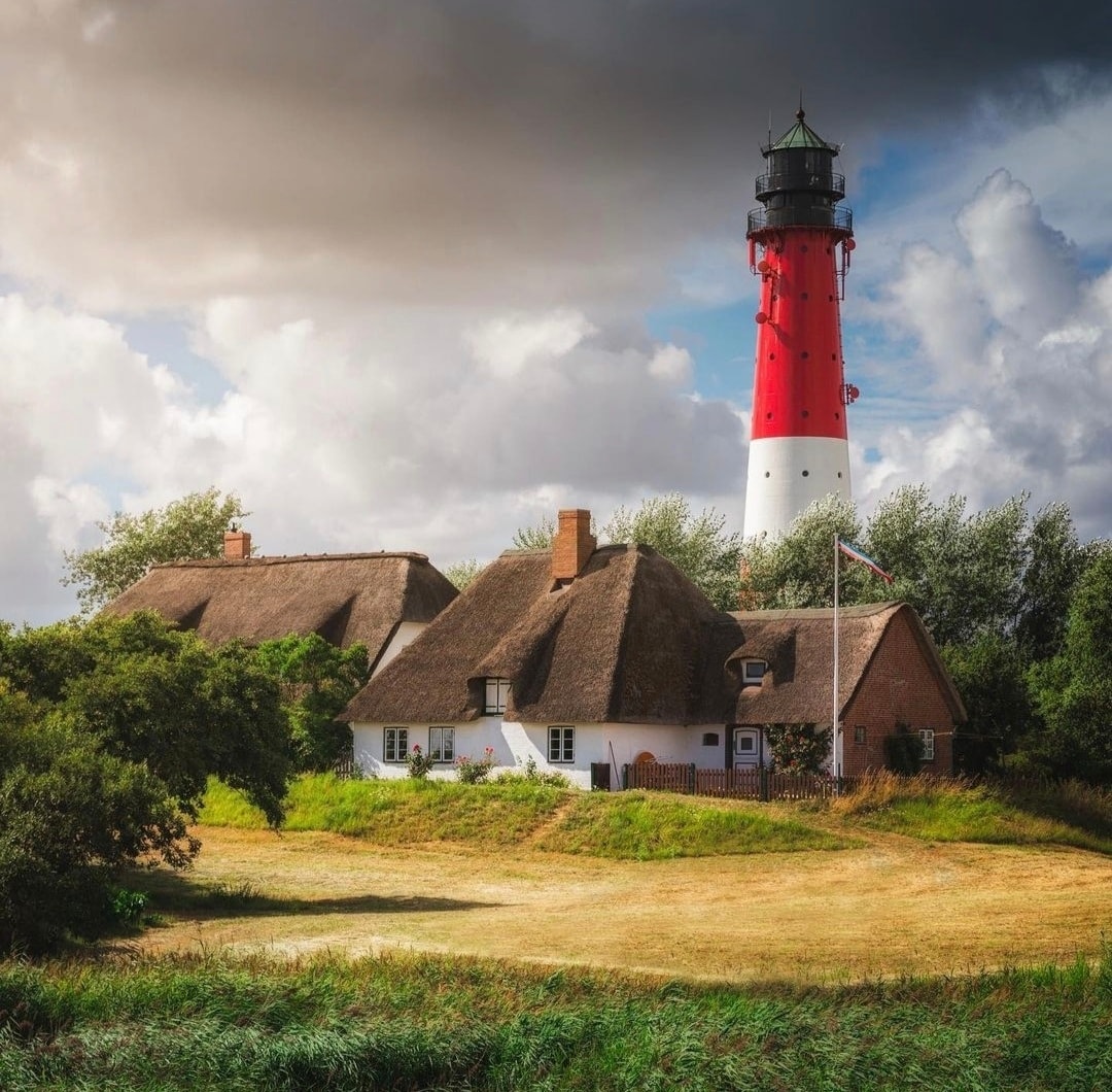 Red and white lighthouse next to thatched cottage under stormy sky