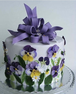 White decorated cake with purple pansies, yellow flowers, green leaves, and purple ribbon bow on top