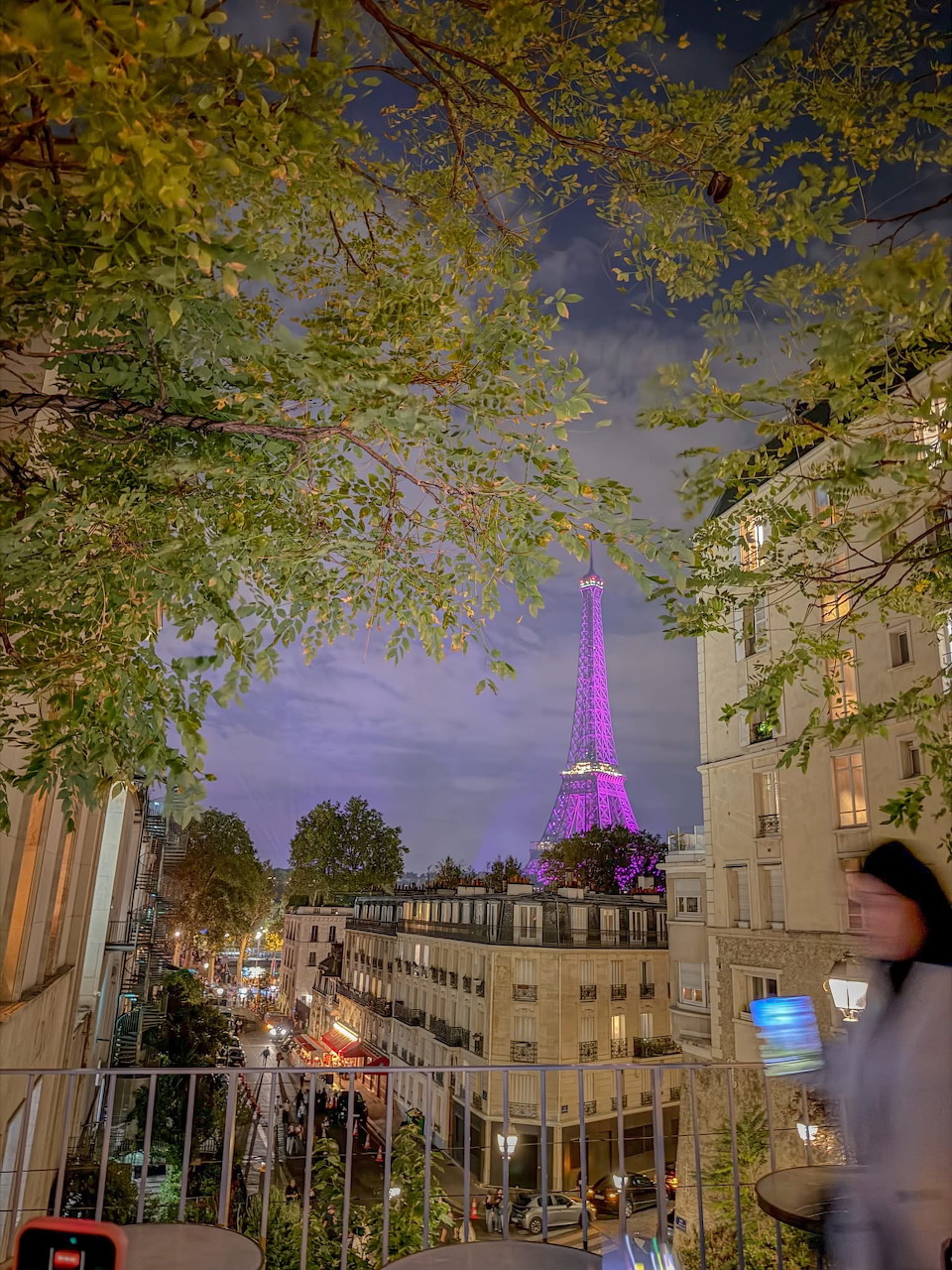 Purple-lit Eiffel Tower beyond Paris buildings, framed by trees with a blurred pedestrian in front.