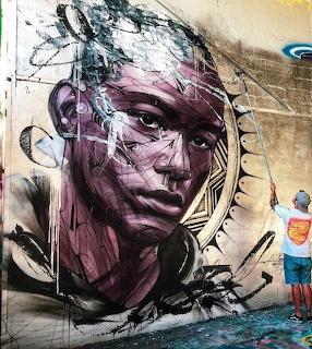 Large street art mural of a woman's face in purple tones with artist painting on wall